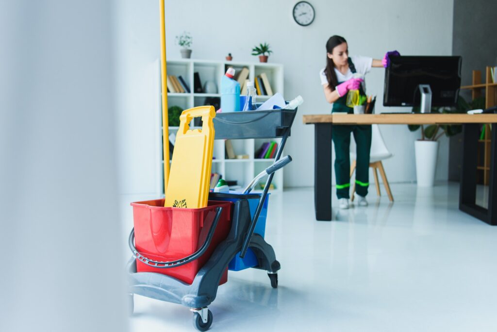 young female janitor cleaning office with various cleaning equipment