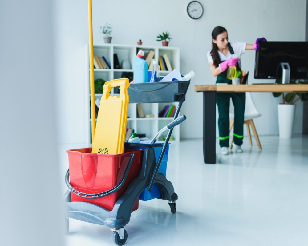 young female janitor cleaning office with various cleaning equipment